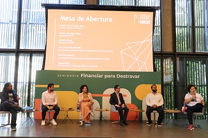 Participantes do evento sentados em frente slide/cartaz de abertura do evento
