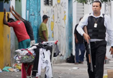 Reprodução de frame do documentário Guarda armado caminha por lugar pobre, com lixo e escombros, e homens virados para a parede