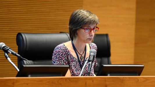 Mulher de cabelo curto e óculos sentada em mesa num palco diante de um laptop