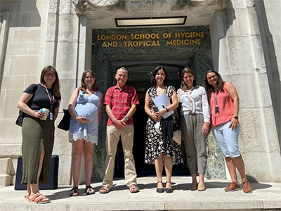 Grupo de pessoas brancas posa para foto em frente à porta da The London School of Hygiene & Tropical Medicine. Da esquerda p/ a direita: mulher de cabelos escuros longos, camisa preta e saia verde, mulher, grávida, de cabelos escuros e longos e vestido azul curto, homem de cabelos curtos grisalhos e camisa vermelha e calça bege, mulher, de cabelos médios escuros e vestido florido, mulher de cabelos médios escuros de camisa branca e calça cinza e mulher de blusa rosa e bermuda jeans.