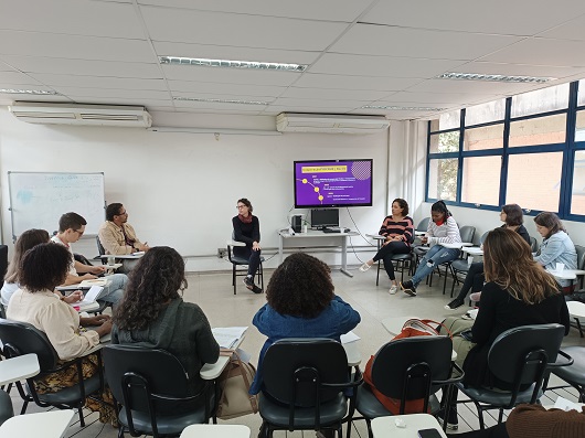 Pesquisadores sentados em roda diante de uma tela, com auxílio da qual uma pesquisadora faz sua apresentação. Sala grande, paredes brancas, janelas amplas.