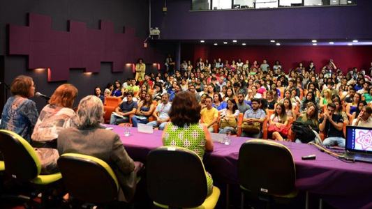 Pessoas sentadas numa mesa num palco, uma delas ao microfone, diante de plateia lotada 