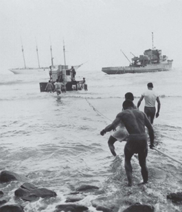 Ao fundo o navio NE Almirante Saldanha, mais à frente uma corveta da Marinha do Brasil e uma balsa sendo puxada por marinheiros em terra através da arrebentação, desembarcando material vindo do Rio de Janeiro