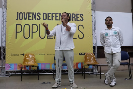 Dois homens negros à frente do banner de lançamento do projeto Jovens Defensores. Um deles fala ao microfone