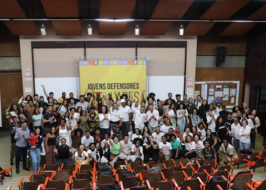 Uma centena de pessoas, de pé e sentadas, no palco de um auditório, acenam para a câmera. Ao fundo, um banner do lançamento do projeto Jovens Defensores Populares