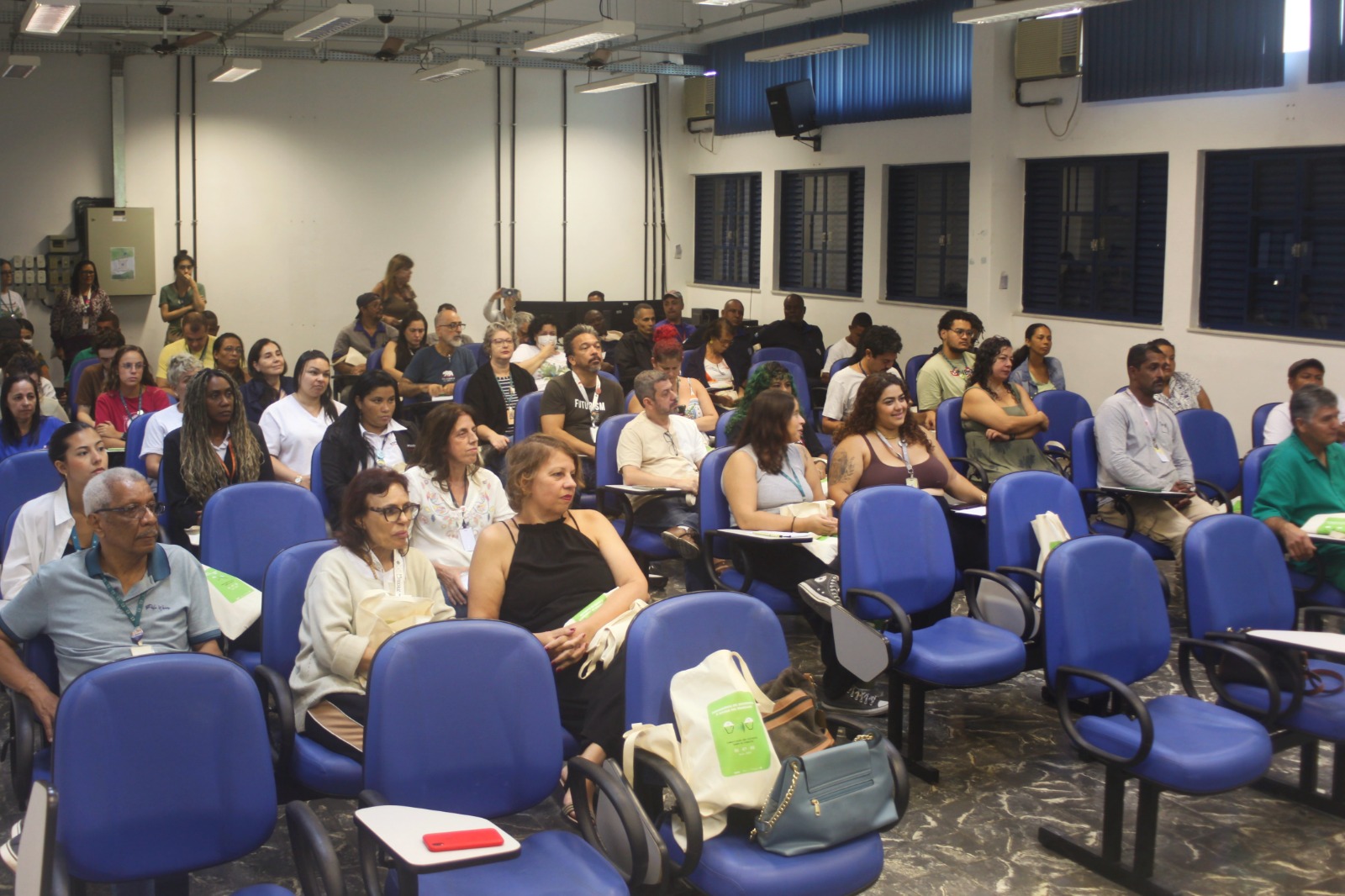 Foto do público do evento. Há cerca de 40 pessoas em uma sala com cadeiras azuis. Ao fundo, algumas pessoas assistem à palestra em pé.