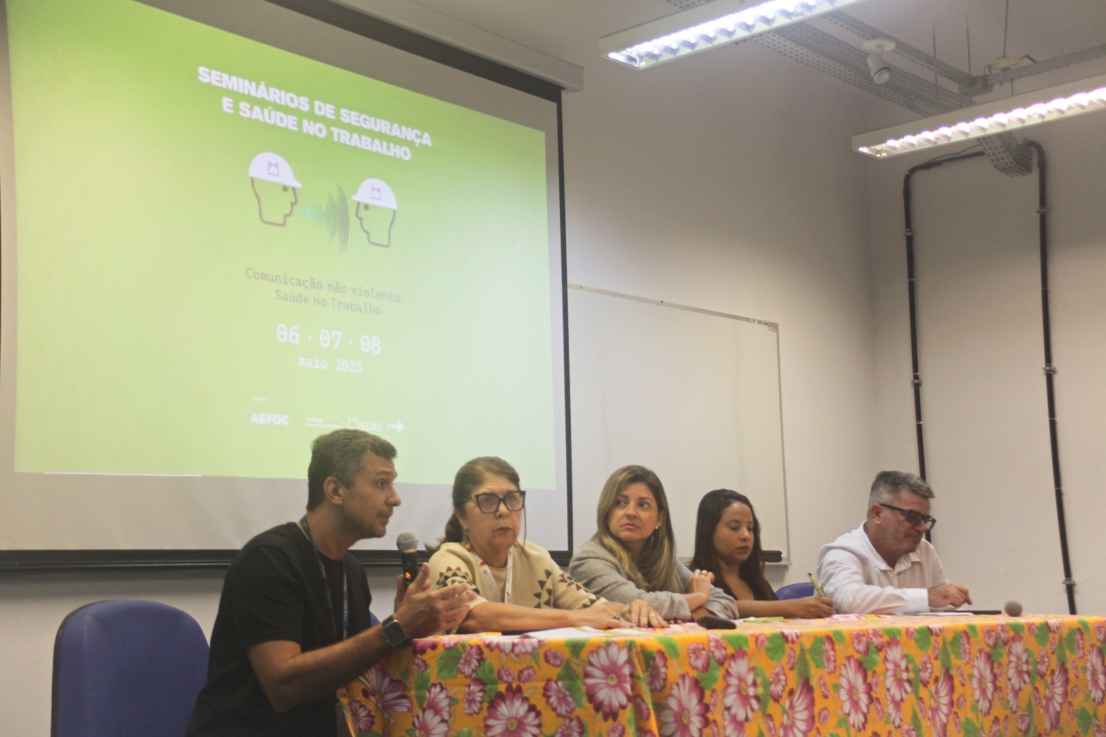 Foto da mesa do evento. Participam cinco pessoas. Nas pontas, estão dois homens brancos. Um deles fala ao microfone. No meio, háduas mulheres brancas e uma mulher negra. Esta última faz anotações.