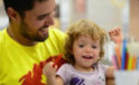 Rapaz de blusa amarela e barba sorri e segura no colo menina com cabelos claros, sorridente e com lápis na mão 