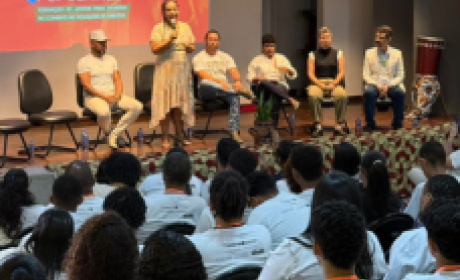 Jovens Defensores Populares chega ao Nordeste com lançamento no Recife jovens assistem mulher falando em palco acompanhada de 5 pessoas sentadas.