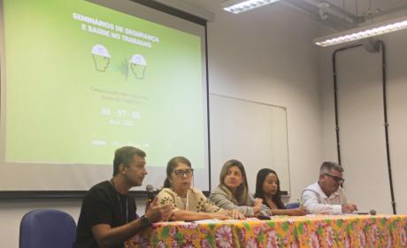 Foto da mesa do evento. Ela é composta de cinco pessoas. Em cada ponta, está sentado um homem. Um deles fala ao microfone. No meio, há duas mulheres brancas e uma mulher negra, que faz anotações.