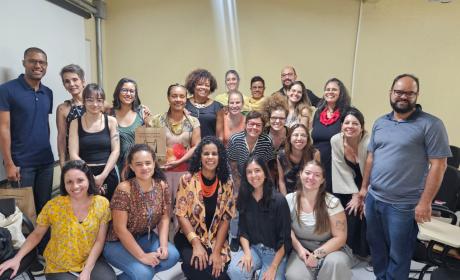 Foto com 15 pessoas em sua diversidade de genero, raça e cor, numa sala amarela. Todas estão sorrindo. 