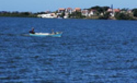Foto: Raul Santana / Multimagens Fiocruz Barco com dois pescadores atravessando um rio, com uma cidade ao fundo