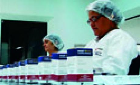 Foto: Divulgação Farmanguinhos Mulheres com uniforme operam a linha de produção, com várias caixas do remédio