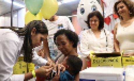 Criança no colo da mãe sendo vacinada por uma profissional de saúde. Ao fundo, está a ministra da Saúde, Nísia Trindade Lima, que observa a cena