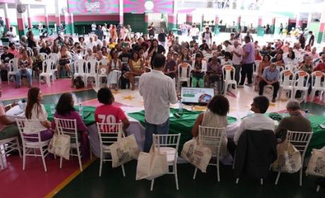 Multidão reunida na quadra da escola de samba da Mangueira. No foco da imagem, um homem branco, usando uma blusa branca, está de pé falando ao microfone, e, ao seu lado, da esquerda para a direita, uma mulher branca, quatro mulheres negras e uma homem branco sentados. Ao fundo, um grande grupo de pessoas assistindo setados em cadeiras brancas de plástico.