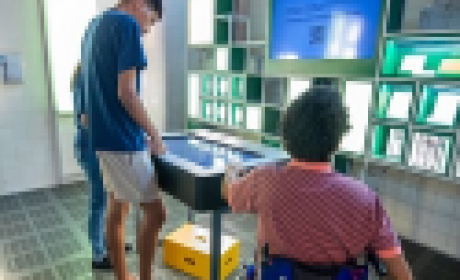 Adolescente e um homem adulto em uma cadeira de rodas visitando uma exposição na Fiocruz 