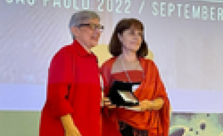 Pesquisadora da Fioruz Bahia recebe prêmio Women in Science Aldina Barral vestindo blazer longo vermelho, recebendo medalha. ao fundo apresentação do evento em roxo com dizeres brancos.
