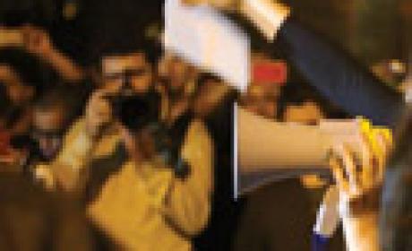Imagem de dentro de um protesto, com mão ao megafone e homem com máquina fotográfica