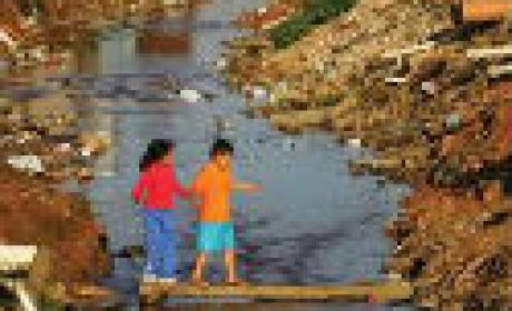Foto: IOC Duas crianças atravessando umaa ponte sob um canal de esgoto a céu aberto