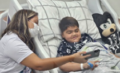 Menino deitado numa cama de hospital e uma enfermeira que mostra o celular para ele 