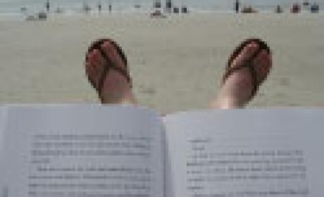 Livro aberto em primeiro plano, com pés em sandálias e paisagem de praia ao fundo