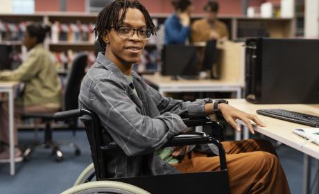 Estudante negro de dreads no cabelo, sentado em uma cadeira de rodas. Ao fundo uma biblioteca